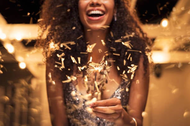 Cheerful young woman holding single sparkler in hand outdoor. Detail of african girl celebrating new year"u2019s eve with bengal light. Closeup of beautiful woman holding a sparkling stick at party night.